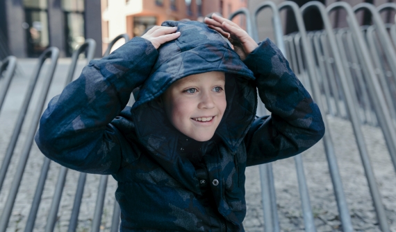 A boy in a patterned blue Pengu KIDS jacket with the hood up
